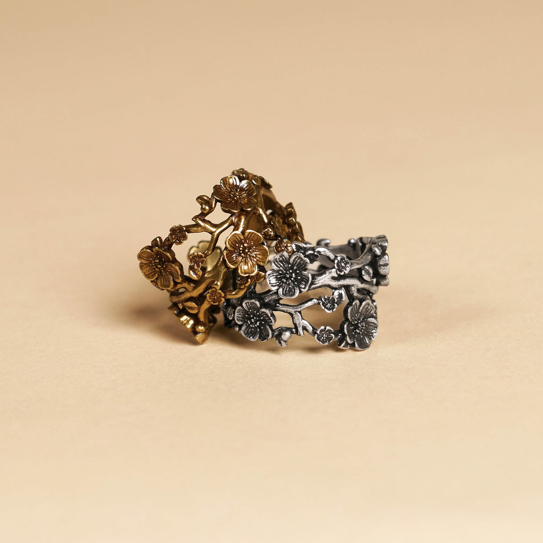 A two-tone metal ring featuring a bronze-toned floral branch design overlaid on a textured silver-toned band, displayed on a warm beige background.