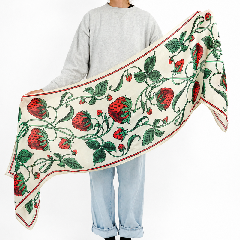A person wearing a light gray sweatshirt, light blue jeans, and black socks holds up a cream-colored scarf with a red and green strawberry vine pattern against a plain white background.